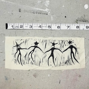 May include: A cream-colored fabric rectangle with a black linocut print of four stylized figures holding hands. Each figure has a star-shaped head. A measuring tape is above the fabric, showing inches.