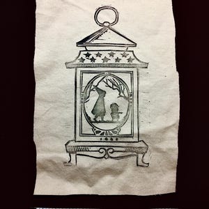 May include: Black and white linocut print of a lantern with a scene inside. The lantern has a decorative top with stars and a ring at the top. The scene inside shows two figures under a tree. The print is on textured paper.