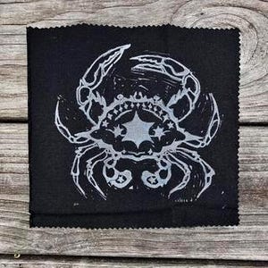 May include: A square, black fabric patch featuring a white linocut print of a crab. The crab design includes a star and crown motif. The patch has a textured appearance and is displayed on a weathered wooden surface.