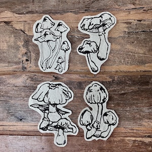 May include: Four handmade mushroom-shaped paper cutouts. Each cutout features a different design of black ink mushroom illustrations. The cutouts are arranged on a wooden surface.