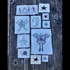 May include: Arrangement of small, light blue fabric squares with black ink illustrations. Designs include stars, a beetle, a snail, a crescent moon, and a butterfly. The squares are arranged on a weathered wooden surface.