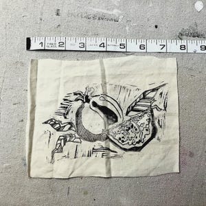May include: A black and white linocut print of a fruit with leaves on a beige fabric. The print is approximately 4 inches wide. A measuring tape is above the fabric.