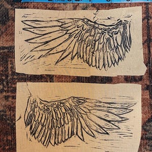 May include: Two black and white linocut prints of a single wing on a brown paper background.
