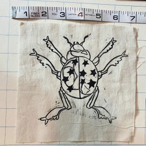 May include: A black and white linocut print of a beetle with star patterns on its back. The print is on a square piece of off-white fabric. A measuring tape is in the background, showing inches.