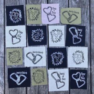 May include: A collection of square fabric patches in black, white, and olive green featuring screen-printed designs. The graphics include interlocking gender symbols, heart-shaped carabiners, and carabiners with text like butch4butch and fem4fem.