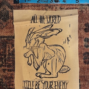 May include: A brown paper print with a black and white illustration of a rabbit. The text "ALL THE WORLD WILL BE YOUR ENEMY" is printed below the rabbit.