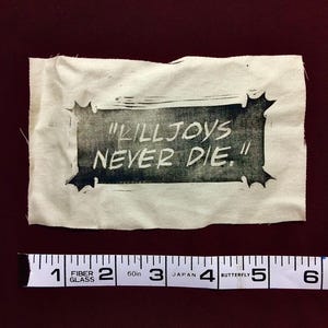 May include: A small, rectangular fabric piece with the text "KILLJOYS NEVER DIE." printed in black. The text is framed by a decorative border. A measuring tape is placed below the fabric, showing inches.