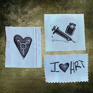 May include: Three small fabric squares with black linocut designs. One features a heart with a transgender symbol, another a syringe and vial, and the last reads "I heart HRT". The designs are printed on white fabric.