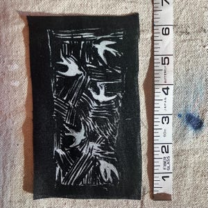 May include: A black fabric patch with a white linocut print of birds in flight. The patch is approximately 4 inches long and 2 inches wide. A measuring tape is next to the patch.