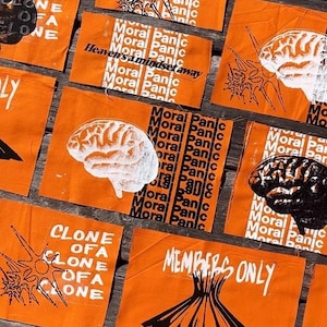 May include: A collection of rectangular orange fabric patches featuring screen-printed graphics. Designs include white and black brain illustrations, the text "Moral Panic," "Clone of a Clone," "Members Only," and "Heaven's a mindset away."