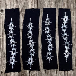 May include: Four black fabric patches with a white chain and spiked ball design. The patches are rectangular with a jagged edge, and the chain design is vertically oriented.