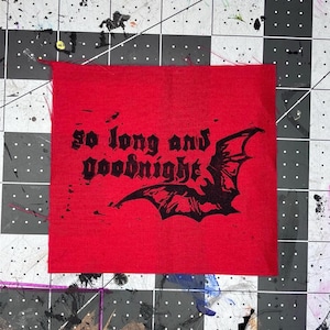 May include: A square red fabric patch featuring a black screen-printed graphic of a bat and the text "so long and goodnight" in a gothic-style font. The fabric has a visible woven texture and raw, unfinished edges on the top and sides.