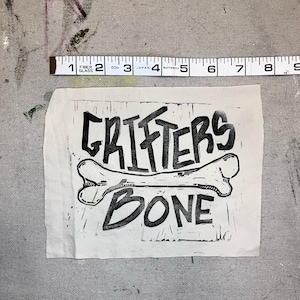 May include: A black and white print on a piece of paper. The print features the words "CRIFTERS BONE" with a bone graphic. A measuring tape is above the print, showing inches.