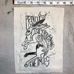 May include: A black and white linocut print on fabric featuring a rabbit with a sword through its head and torso. The text reads "PRINCE OF A THOUSAND ENEMIES". The fabric measures approximately 6 inches wide.