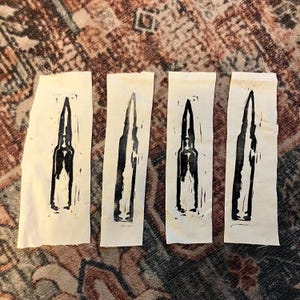 May include: Four rectangular pieces of off-white fabric, each featuring a black linocut print of a bullet. The prints vary slightly in detail and are arranged side by side. The fabric pieces are on a patterned surface.