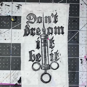 May include: A rectangular white fabric patch featuring a black ink block print of a medical syringe centered vertically. The text "Don't dream it be it" is printed in a gothic-style font, with the words arranged around the syringe illustration.