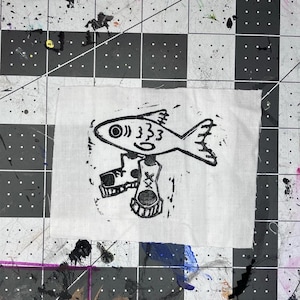 May include: A rectangular piece of white fabric featuring a black ink block print of a fish wearing two high-top sneakers. The fish is depicted in profile with a circular eye and simple fin details, while the shoes show laces and a cross mark.