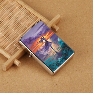 May include: A silver metal lighter with a vibrant image of a dancer on a body of water at sunset. The artwork features shades of purple, orange, and teal, creating a dynamic and artistic design. The lighter is rectangular with rounded edges.