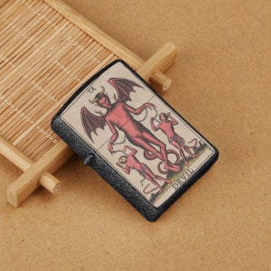 May include: A black lighter with a vintage-style illustration of a red devil with wings and two chained figures. The lighter has the word "DEVIL" printed on it.