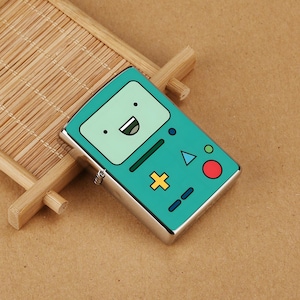 Puede incluir: Un encendedor de color turquesa y plateado con un diseño de personaje de dibujos animados. El encendedor tiene forma rectangular, con un fondo turquesa y un diseño de cara sonriente. Tiene una carcasa de metal plateado y una bisagra lateral.