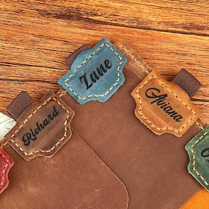 Op de afbeelding: Een bruin leren notitieboek met gekleurde tabbladen met de namen "Daniel", "Richard", "Zane", "Ariana" en "Olivia". De tabbladen zijn respectievelijk rood, bruin, blauw, beige en groen. Het notitieboek is open en toont een oranje binnenkant.