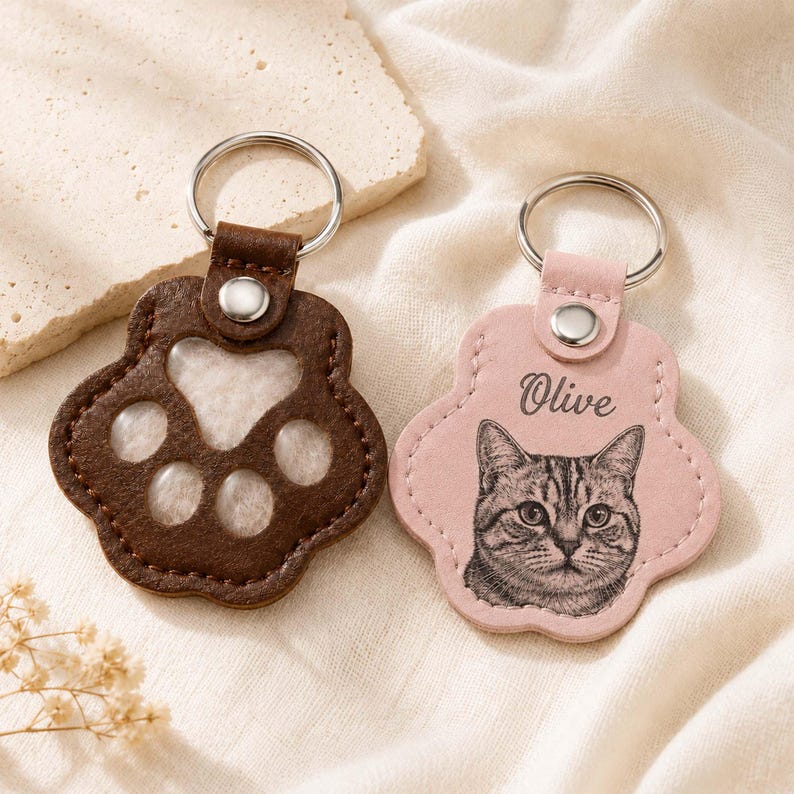 May include: Two paw-shaped keychains with silver metal rings. One is dark brown leather with a cutout paw design. The other is light pink leather featuring a black engraved portrait of a cat and the name Olive printed in a cursive script font.