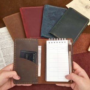 May include: A collection of leather notebooks in various colors, including brown, burgundy, blue, and green. One notebook is open, revealing a notepad and pen. A credit card is visible in a pocket. The notebooks are on a brown surface.