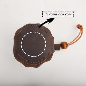 May include: A brown leather item with a circular design, featuring a dotted circle for customization. It has a leather strap with a wooden bead and a knotted end. The text "Customization Zone" is above the item.