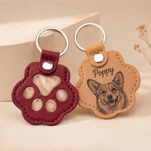 May include: Two paw-shaped keychains with silver metal rings. One is dark red with a cutout center, and the other is tan leather featuring an engraved portrait of a dog and the name Poppy written in cursive script above the dog's head.