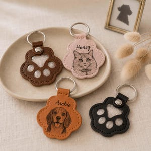 May include: Four paw-shaped keychains with metal rings. Two are solid-colored leather with cut-out paw pads, one is light pink with a cat illustration and the name Honey, and one is tan with a dog illustration and the name Archie engraved.