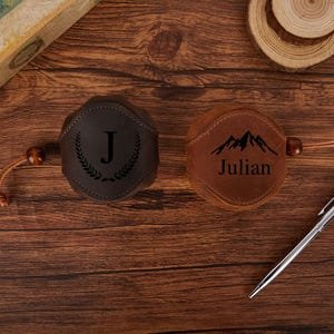 May include: Two leather flask covers, one dark brown with a "J" and laurel wreath design, the other tan with a mountain graphic and the name "Julian". Both have a leather cord and wooden bead detail.