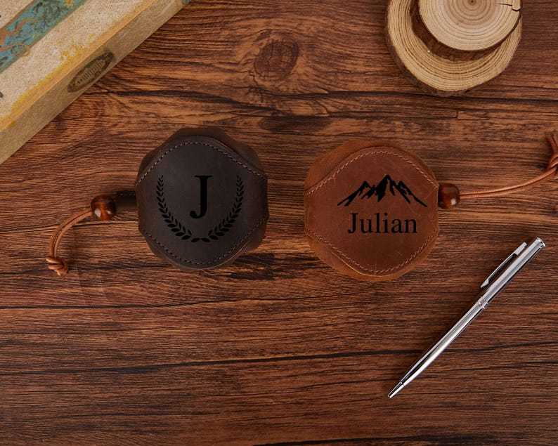 May include: Two leather flask covers, one dark brown with a "J" and laurel wreath design, the other tan with a mountain graphic and the name "Julian". Both have a leather cord and wooden bead detail.