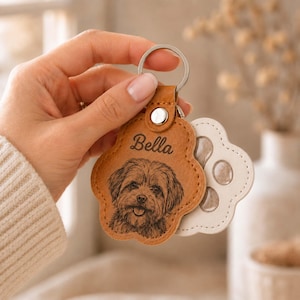 May include: A tan, paw-shaped leather keychain featuring a laser-engraved portrait of a dog and the name Bella in cursive script. The keychain is attached to a silver metal ring and is layered over a white, paw-shaped backing piece.