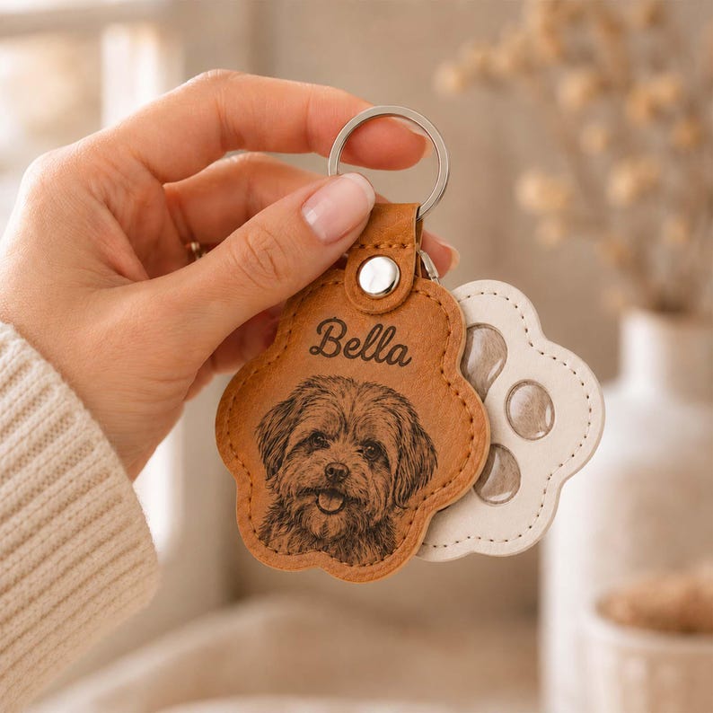 May include: A tan, paw-shaped leather keychain featuring a laser-engraved portrait of a dog and the name Bella in cursive script. The keychain is attached to a silver metal ring and is layered over a white, paw-shaped backing piece.
