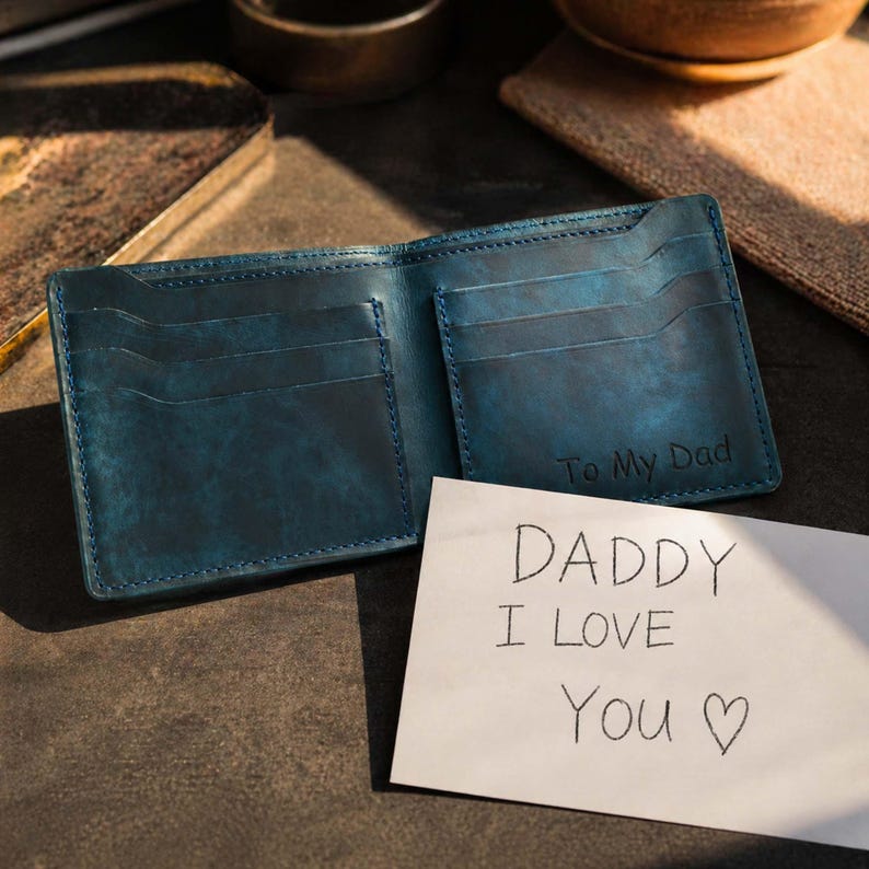 Pu&ograve; includere: Portafoglio in pelle blu aperto, con pi&ugrave; fessure per carte. Le parole "To My Dad" sono impresse all'interno. Un biglietto scritto a mano accanto al portafoglio dice "Daddy I Love You" con un simbolo di cuore.