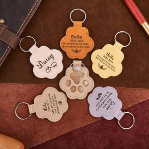 May include: A collection of paw-shaped leather keychains in various colors, including tan, yellow, and lavender. Each keychain has a silver keyring and personalized text, such as names and dates. Some keychains feature a paw print design.