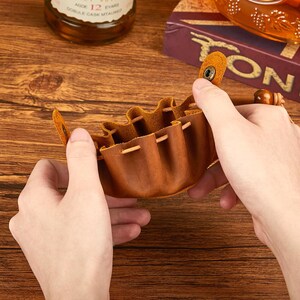May include: A small, brown leather pouch with a drawstring closure and a snap closure. The pouch is held open, revealing the interior. The leather has a textured appearance. The pouch is designed to hold small items.