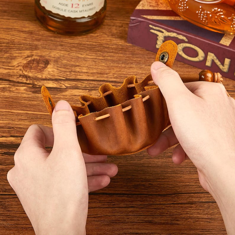 May include: A small, brown leather pouch with a drawstring closure and a snap closure. The pouch is held open, revealing the interior. The leather has a textured appearance. The pouch is designed to hold small items.