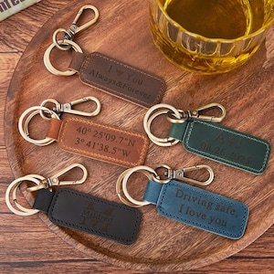 May include: A selection of personalised leather keychains in shades of brown, tan, green, black, and blue. Each keychain has a metal clasp and ring, with engraved text including "I Love You", coordinates, initials, and dates. The keychains are displayed on a wooden tray.
