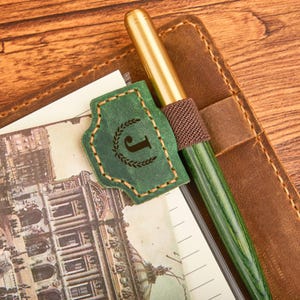 Könnte beinhalten: Ein grüner und goldener Stift liegt auf einem Lederjournal, neben einem grünen Lederlesezeichen mit dem Buchstaben "J" und einem Lorbeerkranz-Design. Das Journal ist auf einer Seite mit einer Vintage-Architekturillustration geöffnet.