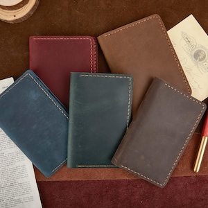 Puede incluir: Varios diarios de cuero en tonos azul, burdeos y marrón. Cada diario está cosido a mano con hilo en contraste. También hay una postal vintage y un bolígrafo rojo y dorado.