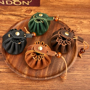 Puede incluir: Una colección de pequeños bolsos redondos de cuero en varios colores: negro, verde y tonos marrones. Cada bolso tiene un cierre de cordón, un adorno de cuentas de madera y un cierre a presión. Los bolsos se exhiben en una bandeja de madera.