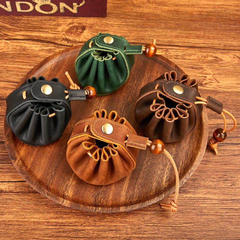 May include: A collection of small, round leather pouches in various colors: black, green, and shades of brown. Each pouch has a drawstring closure, a wooden bead accent, and a snap closure. The pouches are displayed on a wooden tray.