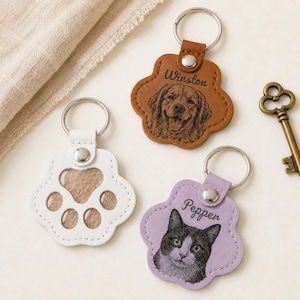 May include: Three paw-shaped keychains with metal rings. One is tan leather with a dog portrait and the name Winston. One is light purple leather with a cat portrait and the name Pepper. One is white leather with cut-out paw pad shapes.