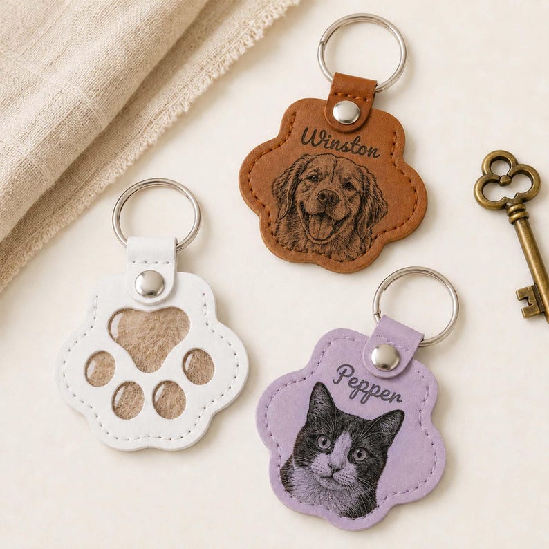May include: Three paw-shaped keychains with metal rings. One is tan leather with a dog portrait and the name Winston. One is light purple leather with a cat portrait and the name Pepper. One is white leather with cut-out paw pad shapes.