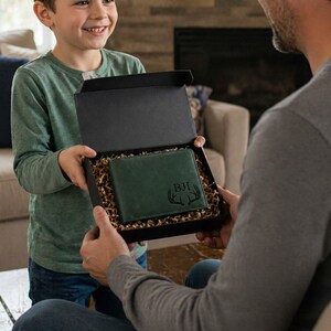 Pu&ograve; includere: Un portafoglio in pelle verde con le iniziali "BJI" e un motivo a corna di cervo &egrave; presentato in una scatola regalo nera riempita di carta sminuzzata. L'immagine suggerisce un regalo, ideale per un'occasione speciale.
