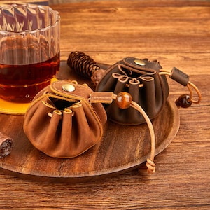 Puede incluir: Dos pequeñas bolsas redondas de cuero en tonos marrones sobre una bandeja de madera. Una es de color marrón claro y la otra es marrón oscuro. Cada bolsa tiene un cierre de cordón y un pequeño adorno de cuentas de madera.