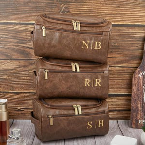 May include: Three brown leather toiletry bags stacked on top of each other, each with a gold zipper and personalized initials. The bags are on a wooden surface, with a wooden bottle opener and small plant in the background.