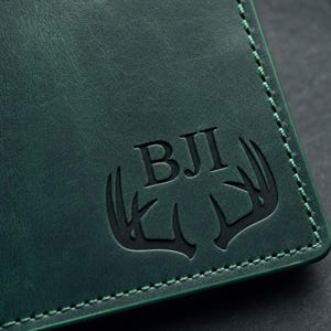 Pu&ograve; includere: Primo piano di un portafoglio in pelle verde scuro con un bordo cucito. Il portafoglio presenta le iniziali "BJI" e un design a corna di cervo in rilievo sul davanti. La pelle ha un aspetto strutturato.