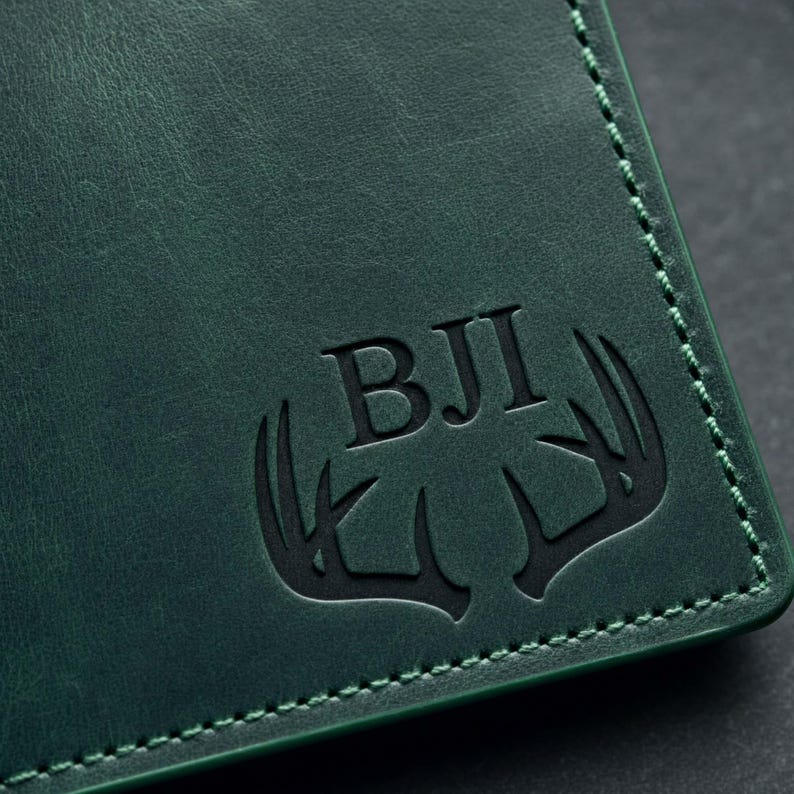 Pu&ograve; includere: Primo piano di un portafoglio in pelle verde scuro con un bordo cucito. Il portafoglio presenta le iniziali "BJI" e un design a corna di cervo in rilievo sul davanti. La pelle ha un aspetto strutturato.